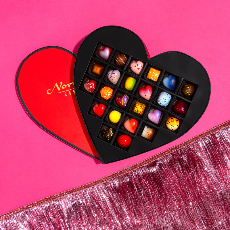 24 beautifully crafted chocolates in a red, heart-shaped gift box. The gift box includes 10 limited -edition hearts as well as 14 signature pieces. The box is offset on a purple background with a festive pink fringe ribbon in the bottom right corner. 